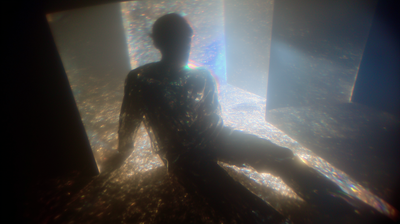 A dark silhouetted figure sits leaning back between large translucent rectangular panels, prismatic light refracting through its glass-like body, scattered chromatic aberration creating rainbow edges across the surface.